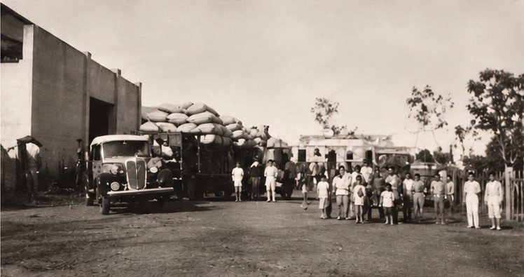 Fotografia histórica em sépia: caminhões carregados de sacas de café e trabalhadores em frente a um armazém