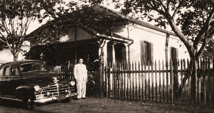 Fotografia histórica em sépia: homem de terno claro ao lado de um automóvel antigo, em frente à casa que abrigava o escritório da Empresa Yanase